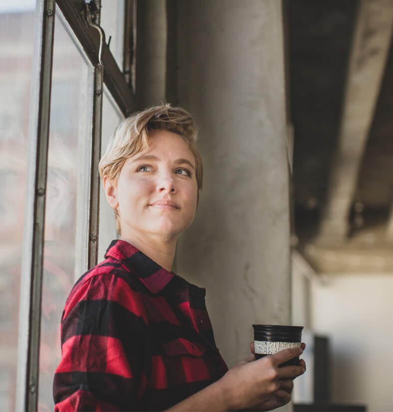 Eine junge Frau steht an einem Fenster, hält einen Kaffee in der Hand und blickt positiv nach vorne.